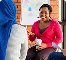 Lady holding a cup of coffee while talking to another lady whose back of head is in shot